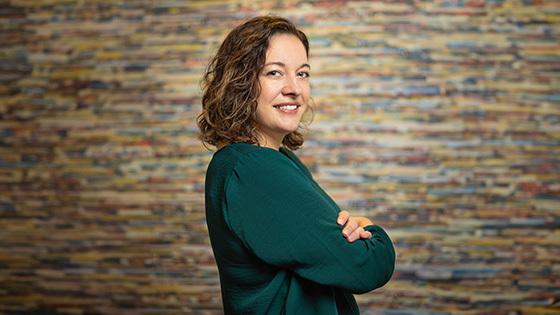 Aron Gelineau stands with her arms crossed and smiles at the camera. She has brown, curly hair and is wearing a teal sweater.